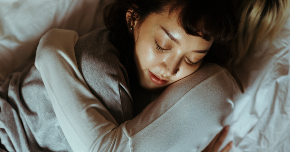 Two people cuddle in bed. Their arms are tightly wrapped around each other and you can only see the face of one of the people who looks calm and content.
