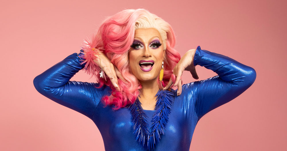 Promo image of Panti Bliss for 'If These Wigs Could Talk'. The photo shows Panti wearing a sparkly Blue dress in front of a salmon coloured background. She is wearing a blonde and pink wig.