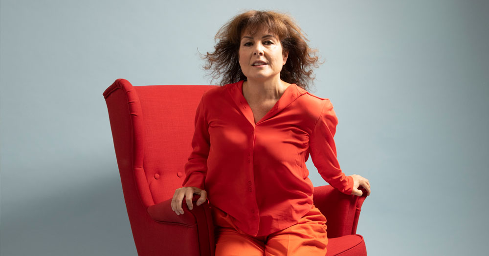 A promotional image for the play Haunted. In the image Tara Flynn is sitting on an orange armchair wearing an orange jumpsuit. She is sitting foward with her hair blowing in the wind.