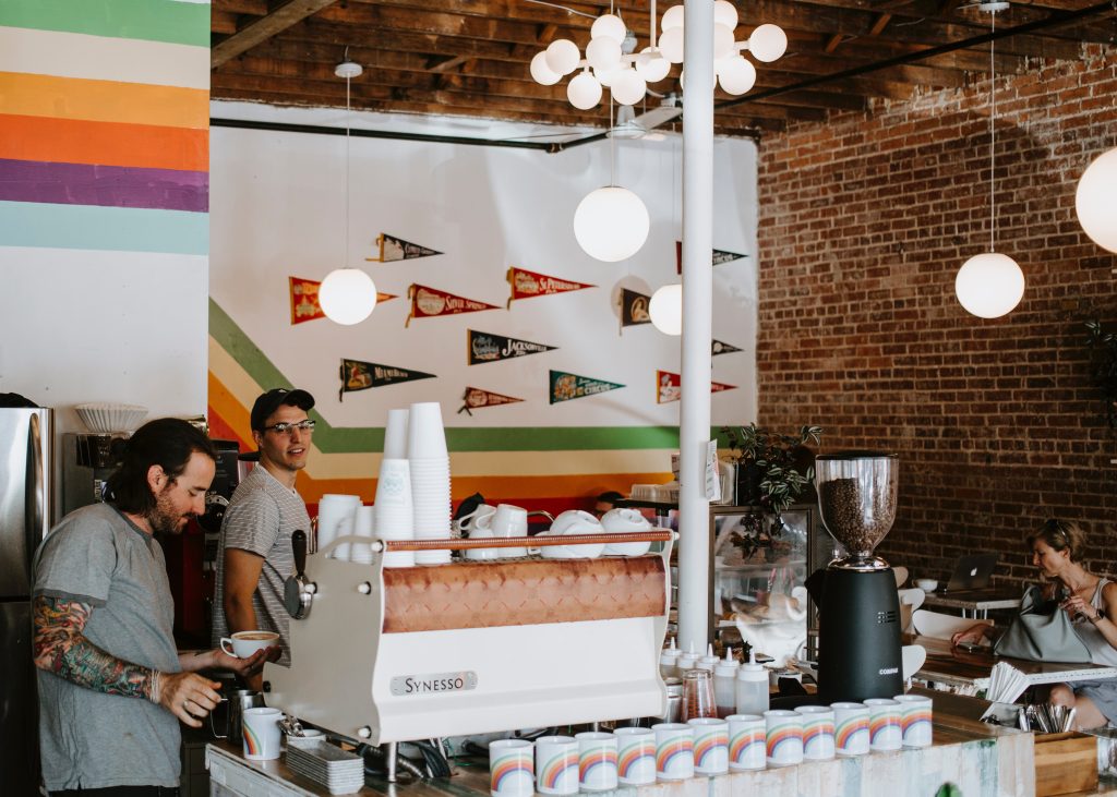 Two baristas standing in a coffee shop with rainbow decor. This piece is forecasting the closure of LGBTQ+ venues.