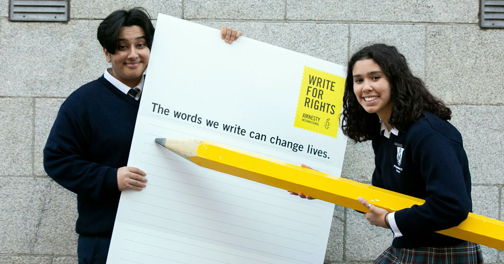 Two students taking part in the Write for Rights campaign, holding a giant piece of paper and a giant pencil.