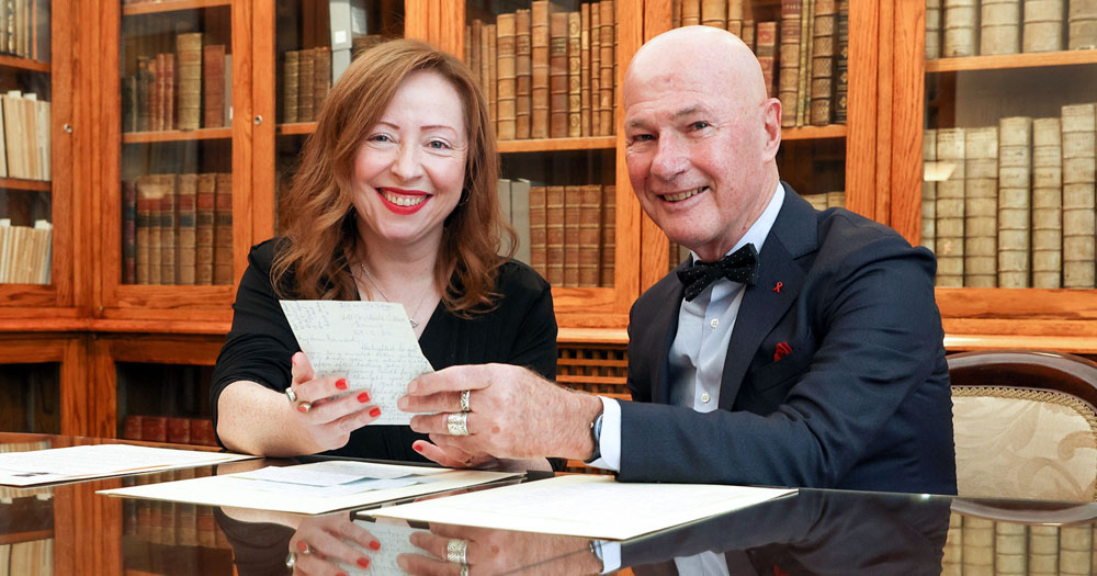 The photograph shows Katherine McSharry of the National Library of Ireland and Bernárd Lynch sitting at a table in a library holding a piece of paper. They are both looking into the camera smiling.