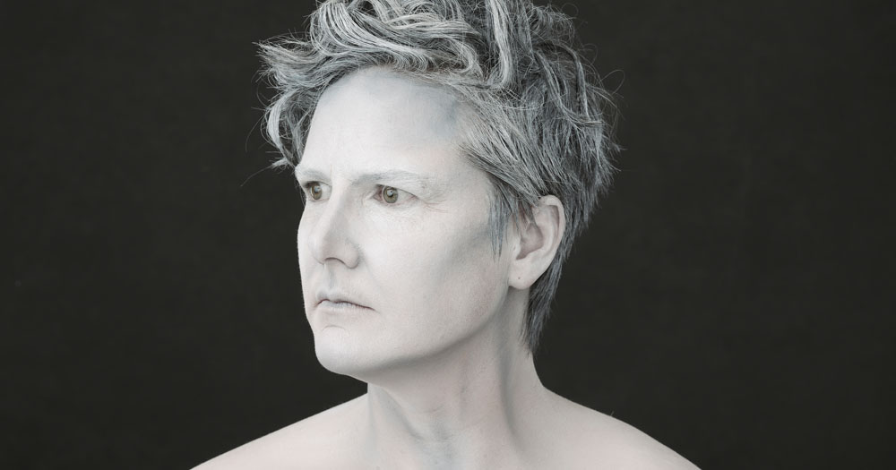 The photograph shows a headshot of Hannah Gadsby. She is facing left and is topless. Her body, face and hair are all painted grey.