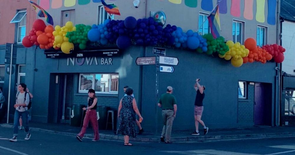Multiple people walk by Galway's only gay bar, Bar Nova, which is painted grey and decorated with many balloons in all the colours of the rainbow.