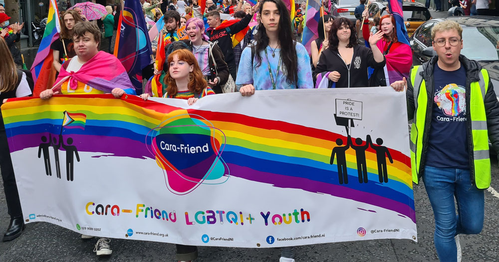 Members of Cara Friend marching in a Pride parade. Everyone is wearing colourful clothing and 5 people are carrying a banner with the words "Cara-Friend LGBTQI+ Youth".