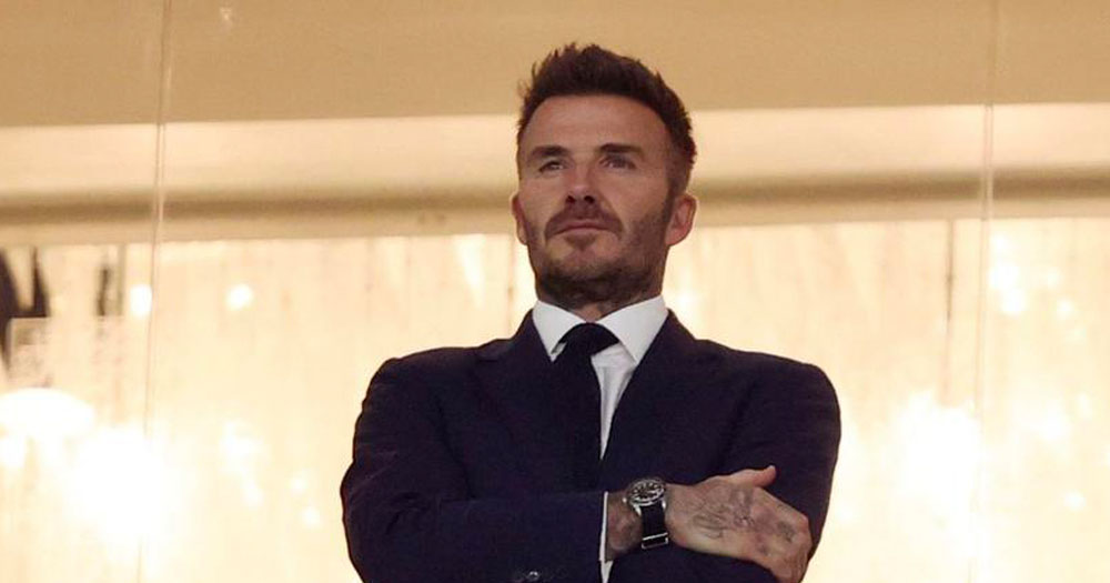 The image shows David Beckham standing with his arms folded as he watches a football match during the Qatar World Cup.