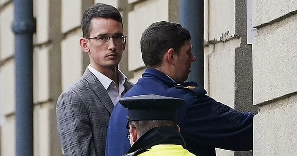 The image shows Enoch Burke, who recently refused to be released for Christmas, being escorted into a building by two Gardaí. He is wearing a grey plaid suit with a white shirt and open colar.