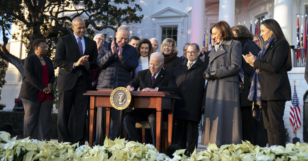 US President Biden signs an act to protect marriage equality, while other politicians applaud and smile.