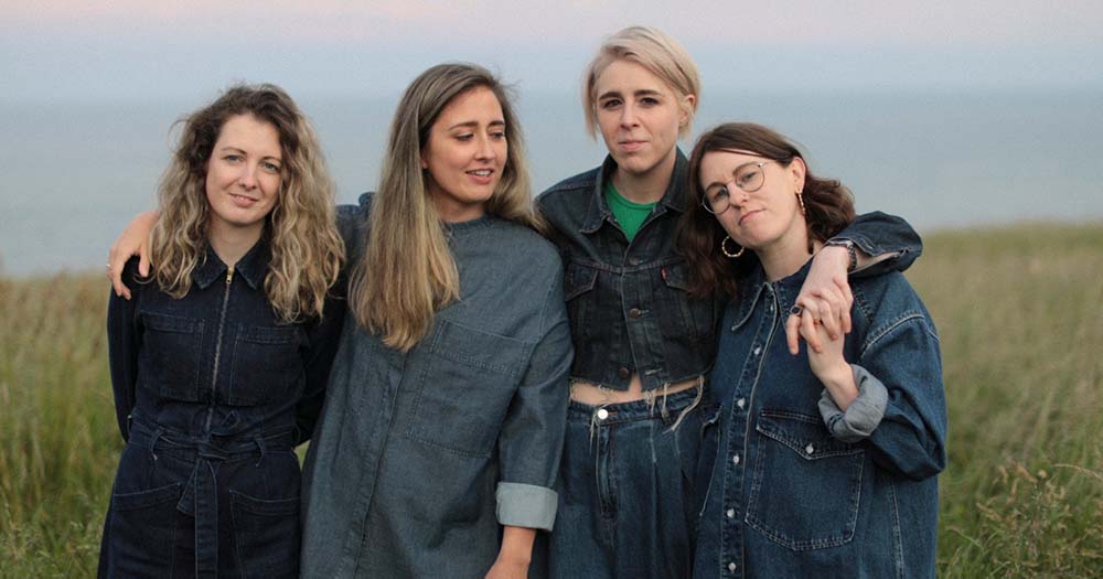 Four members of the iconic queer Irish band, Pillow Queens, stand arm in arm in a field. They will be headlining the annual St. Patrick’s Festival in March 2023!