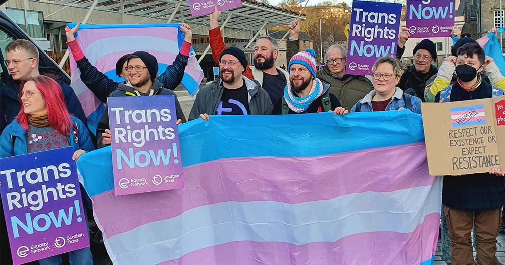 A group of trans rights supporters wearing pink, white, and blue stand with a trans flag in support of Scotland passing their landmark Gender Recognition Reform legislation.