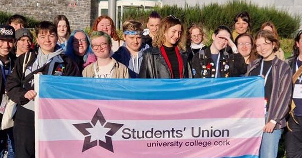 Students from UCC lined up in front of a trans pride flag with Students' Union logo.