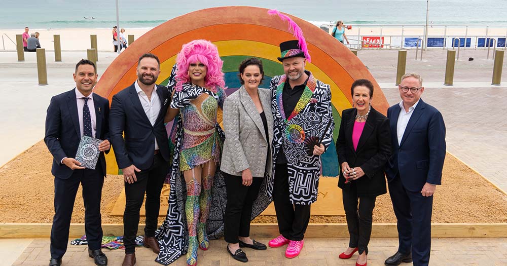 Several people in queer outfits pose in front of rainbow walkway in preparation for WorldPride in Sydney 2023.