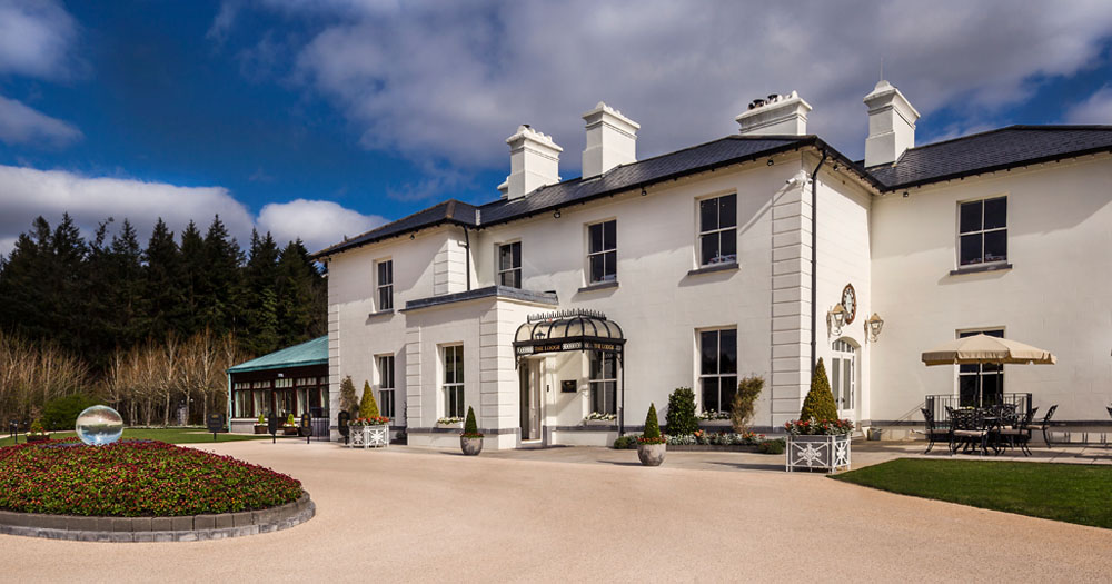 The image shows the front of the Lodge at Ashford Castle. The house is a large white washed country house with a sandy coloured driveway in front.