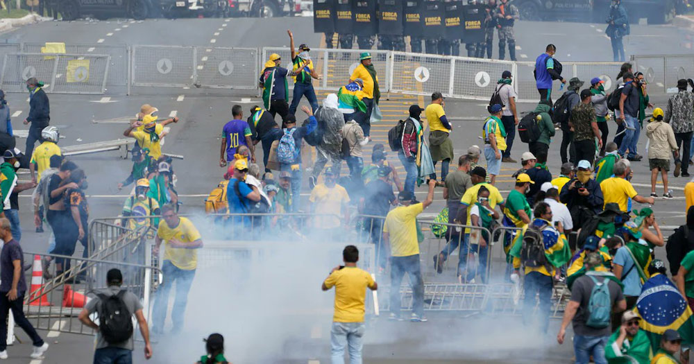 Far-right extremists attacking Brazil's congress. They are dressed in yellow and green, there is smoke and the police is behind barricades.