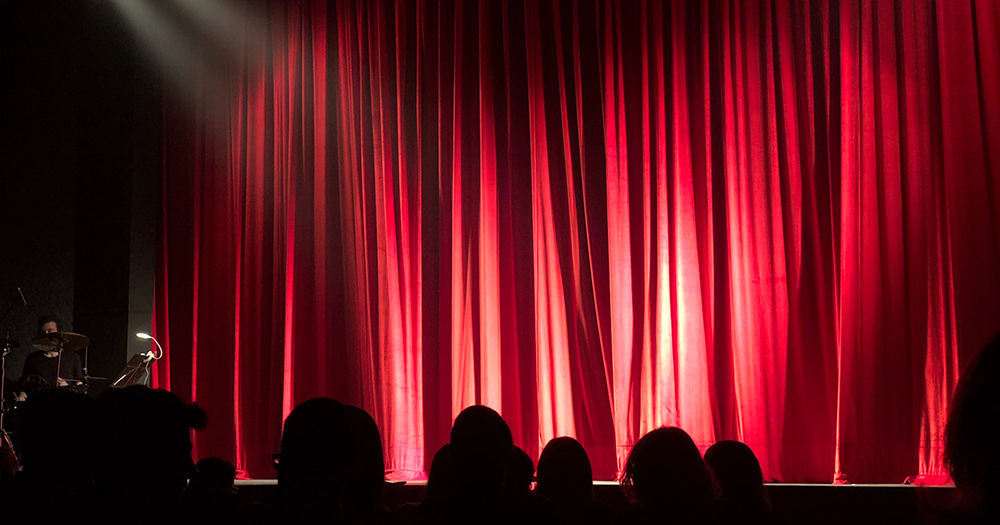 Silhouettes of audience members facing a red curtain because GCN is giving away Candlelight Cabaret tickets.