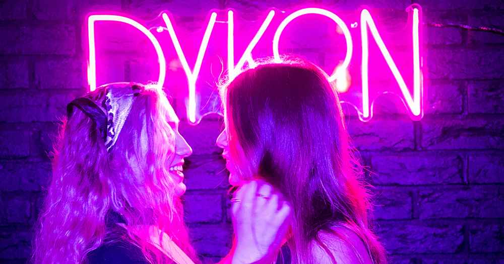 Two girls looking at each other in front of the Dykon sign.