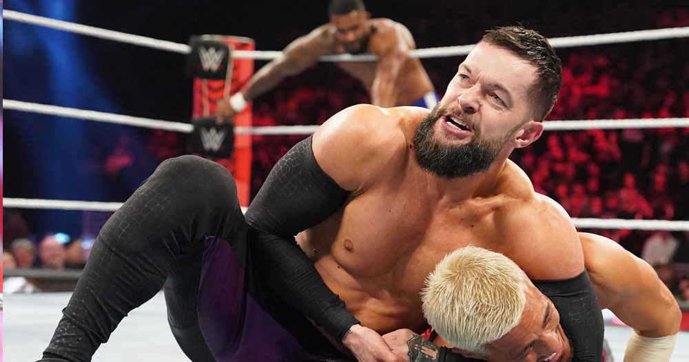 The image shows wrestler Finn Bálor on the ground, leaning over another wrestler whom he has in a headlock.