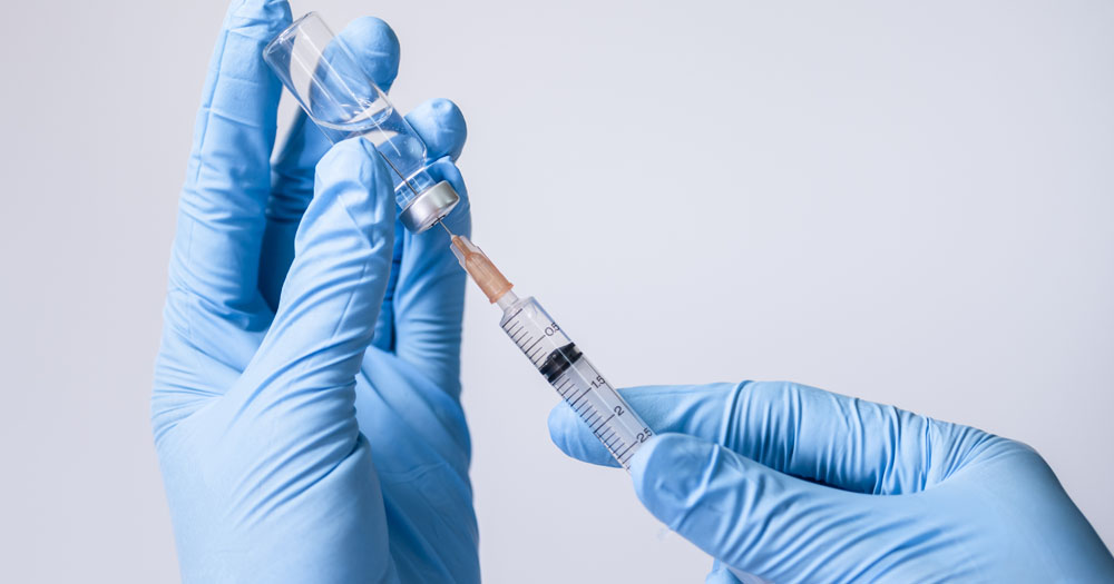 Johnson and Johnson have announced that trials of their HIV Vaccine have been unsuccessful. The photo shows two hands wearing blue latex gloves holding a syringe being stuck into a vial.