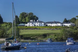 An image of the Lodge at Ashford Castle taken from Lough Corrib. In the foreground there are two boats. The Lodge sits on a hilltop overlooking the lake.