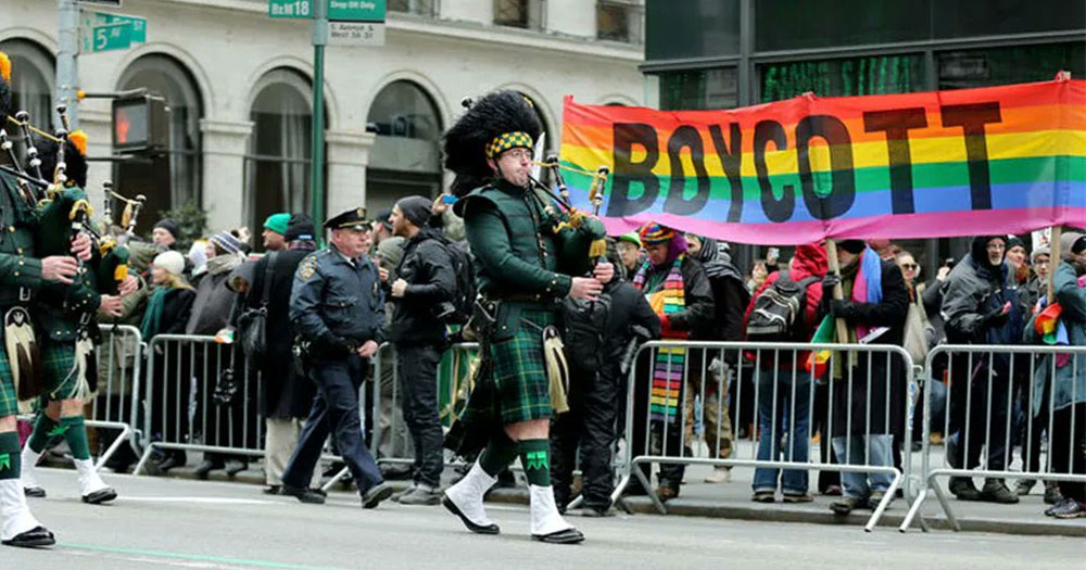 People marching in the Staten Island St. Patrick’s Day Parade, with LGBTQ+ groups protesting on the said with a Pride flag with the word "boycott" printed on it.