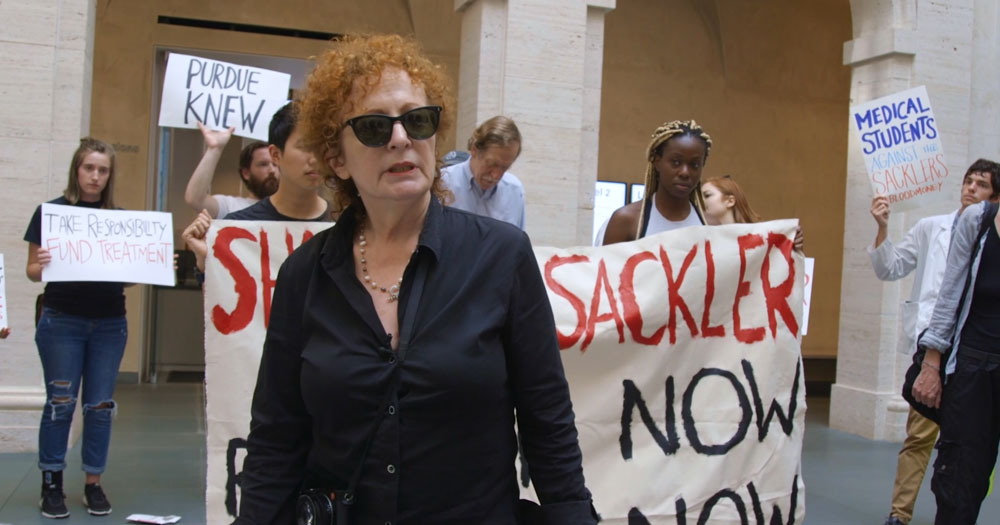 A screenshot from All the Beauty and the Bloodshed starring artist Nan Goldin. The image shows Goldin standing in front of a hand painted banner calling for an end to donations from the Sackler family.
