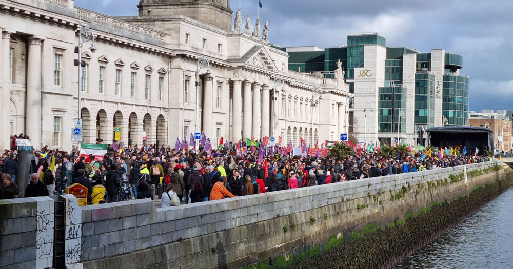Huge crowd participating in solidarity march standing in front of Custom House in Dublin to listen to speeches from speakers on a stage.