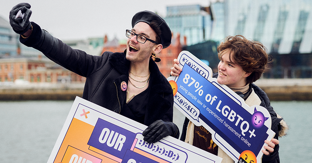 Two LGBTQ+ youth take selfie while holding signs launching social media campaign.