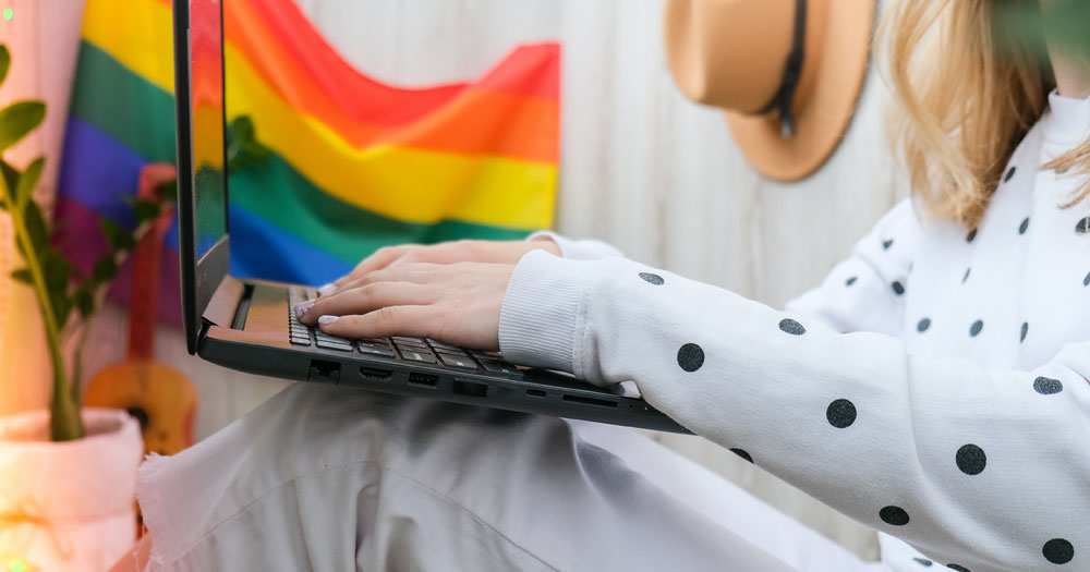 This article is about GCN 2023 Community Feedback Survey. In the photo, a person holding a laptop with a Pride flag in the background.