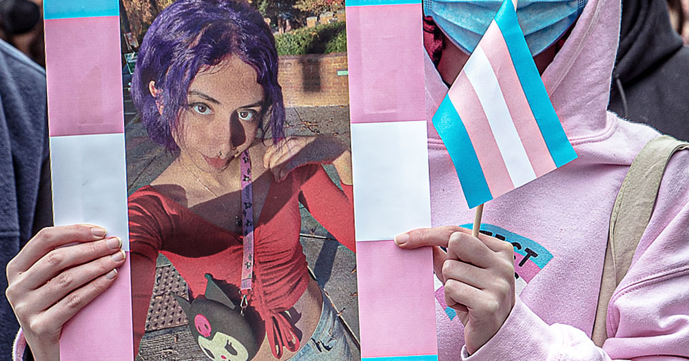 A person holding a photo of Saudi woman Eden Knight at a protest, with a trans flag on one hand.