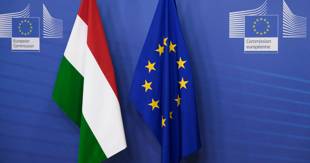 This article is about Ireland joining the case against Hungary's anti-LGBTQ+ law. In the photo, a EU flag and a Hungarian flag in front of a blue background with the logo of the European Commission.