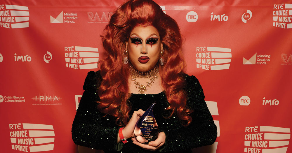 Drag queen Lavender holding the award won by CMAT for best Irish album of the year, while standing in front of a red background.