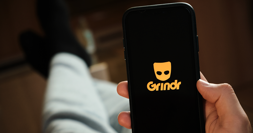 A queer person holding a phone with Grindr on the screen.