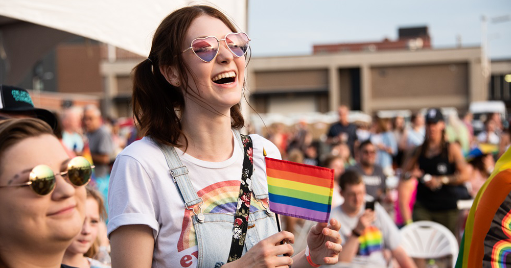 Woman in a crowd wearing sunglasses and carrying Pride flag representing Spartacus Gay Travel guide.