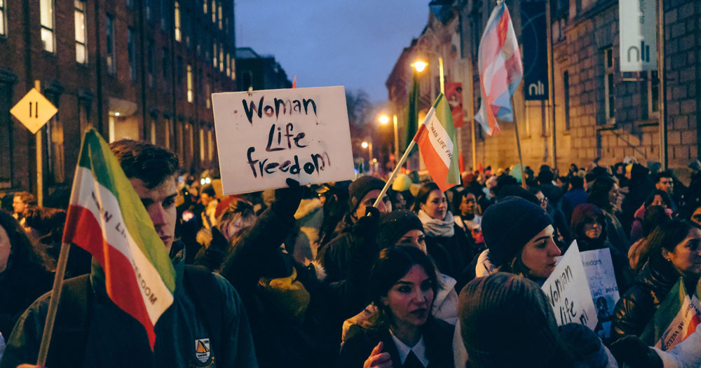 A crowd marching in Dublin on International Women's Day, carrying trans and Iranian flags and signs that read "woman, life, freedom".