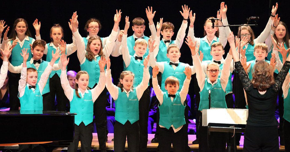 An image of Cór na nÓg who will perform Considering Matthew Shepard alongside Cór Linn. The image shows three lines of children wearing turquoise waistcoats with their arms raised in the air as they sing.