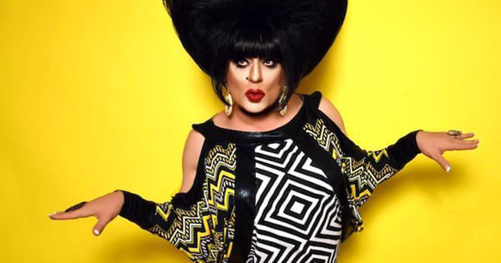 Heklina posing wearing a black wig in front of a yellow background.