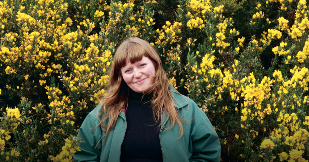 This article is about a residency on climate change for LGBTQ+ people. In the photo, a woman stands in front of yellow flowers in honour of the Homeworks Project.