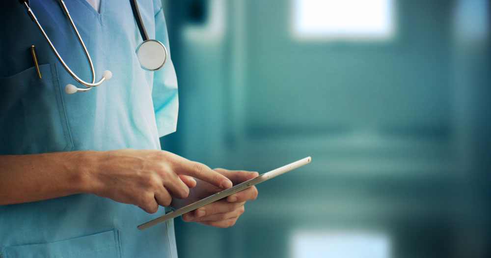 The HSE has published a report on the impact of the closure of the Tavistock clinic on Irish trans patients. The image shows a mid-view of a doctor looking at an iPad. The head and legs are cropped, but he is wearing blue scrubs and has a stethoscope around his neck.