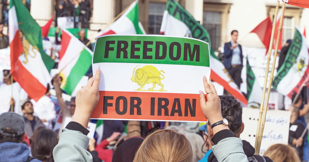 This post is about the LGBTQ+ community in Iran. In the photo, a person holding a banner with the Iranian flag and a message that reads "Freedom for Iran" with people waving Iranian flags in the background.