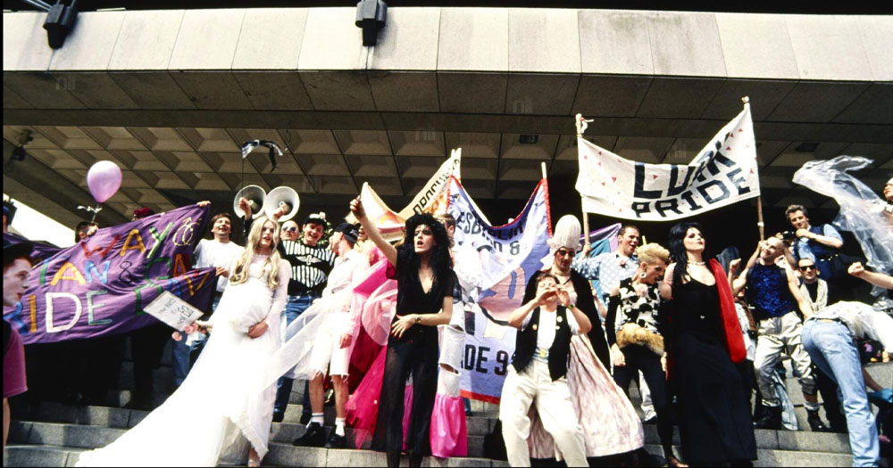 June 2023 markes the 30th anniversary of decriminalisation in Ireland. The photo was taken at Dublin Pride 1993 following the verdict. In the image a crowd are gathered on the steps of a brutalist concrete building (the old Central Bank on Dame Street, Dublin). There is a drag queen at the front with arm raised in a salute. Behind her the crowd are holding up banners from various Pride organisations.