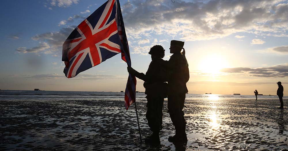 Two British soldiers stand on the edge of sea holding the British flag.