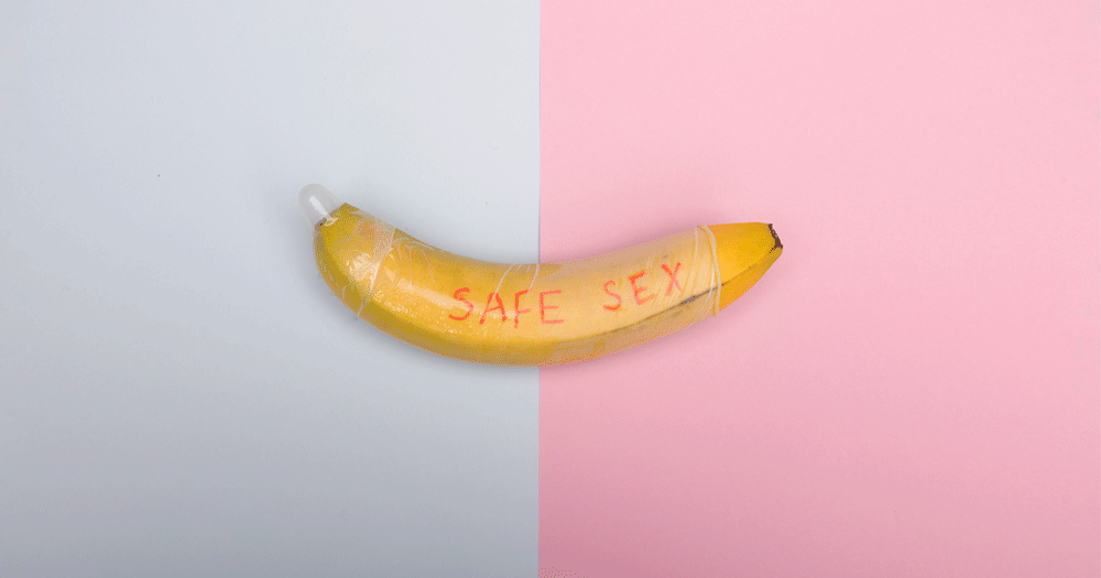A banana wrapped in a condom with the message "safe sex" written on it in red, with a baby blue and baby pink background.
