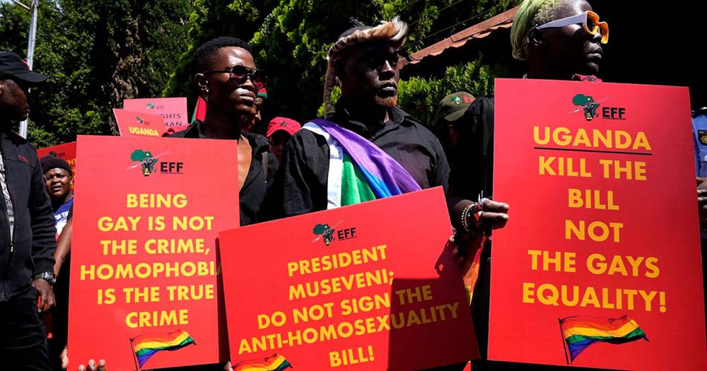 Three people hold signs protesting the Ugandan anti-LGBTQ+ law