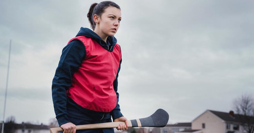 The image is a screenshot from the film Who We Love. It shows a young woman holding a hurl, wearing a red sports bib. She has an angry expression on her face.