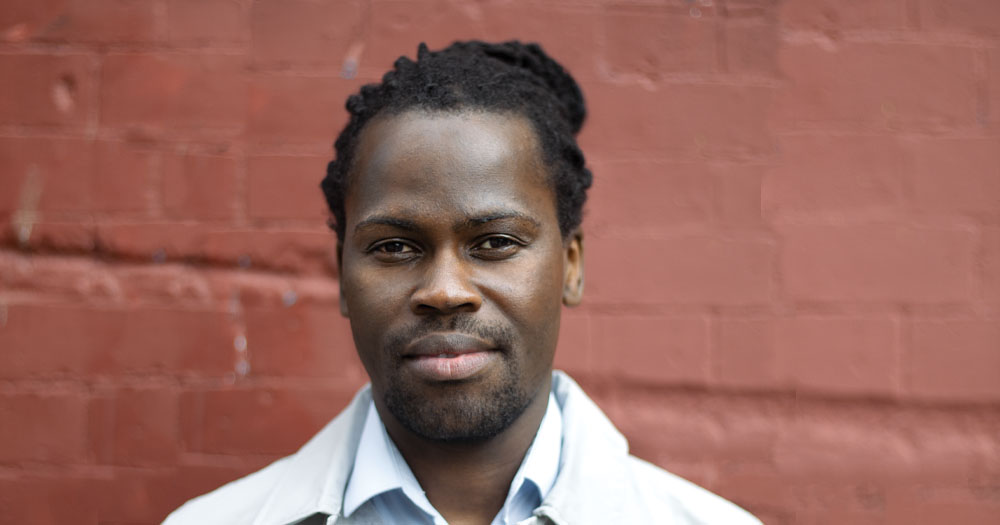 The image is a headshot of MASI activist Bulelani Mfaco. He is standing in front of a red wall wearing a white jacket and light blue shirt. He has long hair tied in a bun.