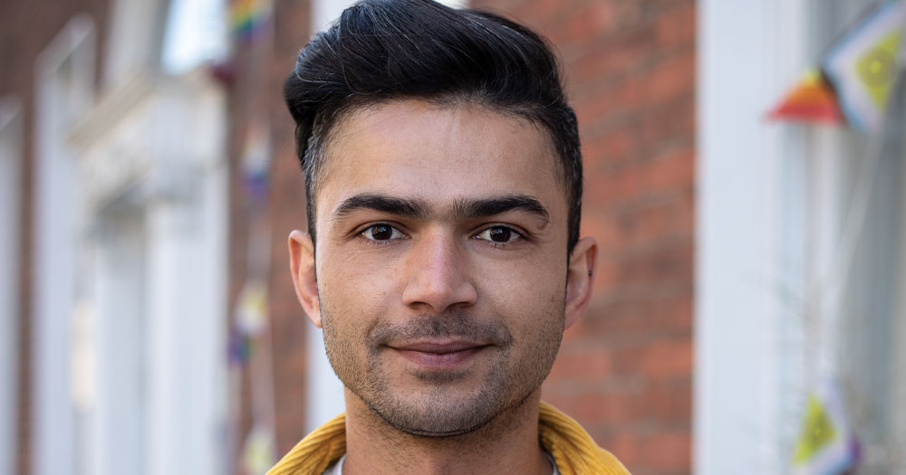 The photo shows Diego Caixeta. It is a close up headshot taken in front of a building with a Pride flag. He is wearing a mustard coloured shirt open over a white tshirt.