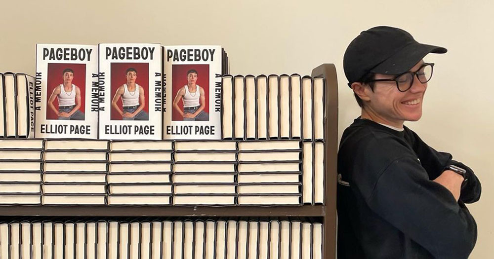The image shows actor Elliot Page leaning with his back against a bookshelf. On the are copies of his new book Pageboy. He is wearing a black baseball cap, glasses and a black sweater. He is smiling with his eyes closed facing away from the camera.