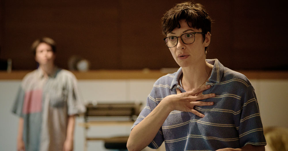 The photograph shows a rehearsal for Fun Home by Alison Bechdel. In the foreground of the image is actor Frances McNamee, who plays older Alison and in the background is Orla Scally who plays middle Alison. Frances has cropped hair and is wearing glasses. She has her hand placed to her chest with a quizzical expression.