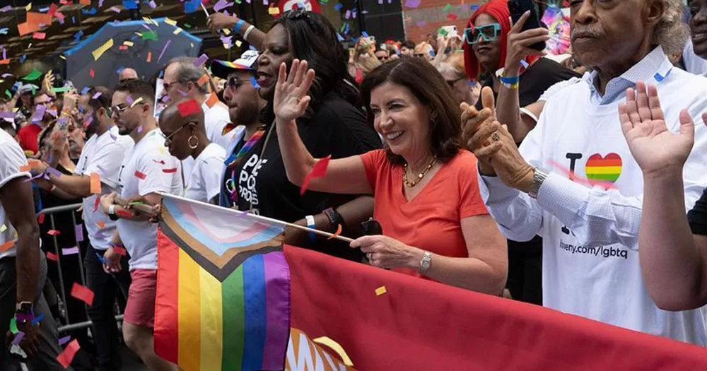 Governor of New York Kathy Hochul waves as she marches in the Manhattan Pride parade. She is wearing an orange v-neck t-shirt and is holding a progressive Pride flag in her other hand.