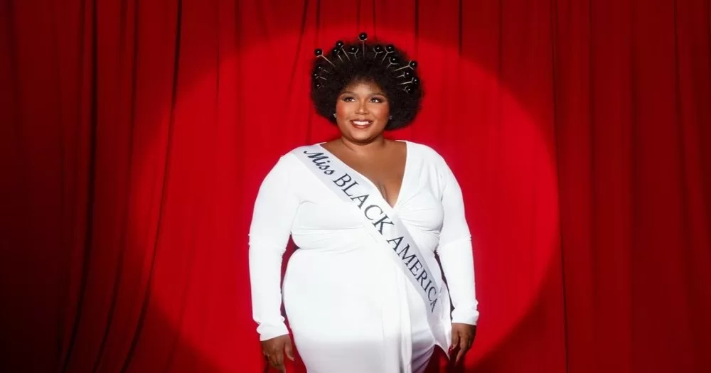 This article is about Lizzo donating money for Black trans women. In the photo, Lizzo in a white dress wearing a shash reading "Miss Black America" against a red backdrop.